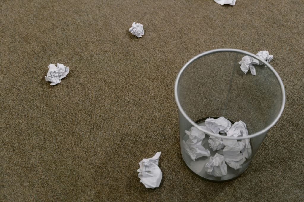 Waste paper basket on the carpet with crumpled up pieces of paper around it.